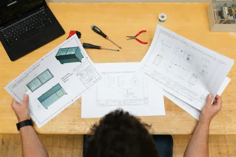 Man reviewing detailed technical blueprints and 3D design drawings at a wooden desk with tools and laptop