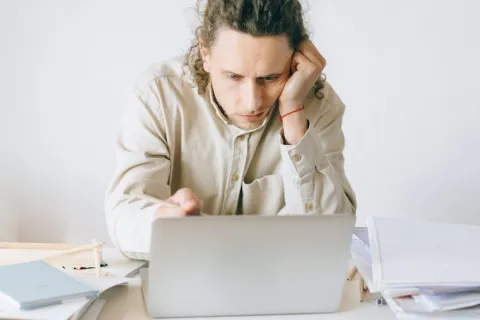 Man with curly hair focused on laptop screen while sitting at desk with notebooks and papers