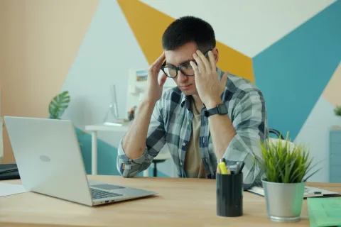 Young man wearing glasses stressed while working on laptop in a modern office with colorful geometric wall art.