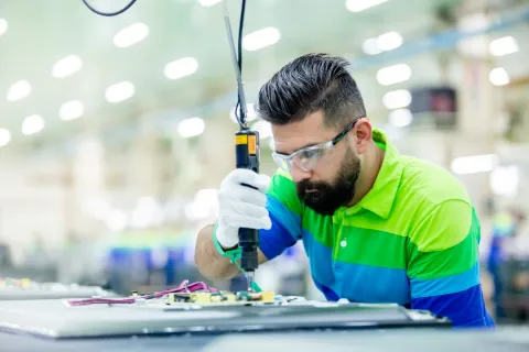 Technician wearing safety goggles using a power screwdriver to assemble electronic components in a factory.