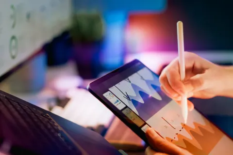 Person using a stylus on a tablet displaying colorful charts next to a laptop in a workspace.