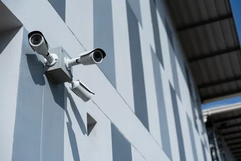 Three security cameras mounted on a blue building wall under a roof watching multiple directions during daylight.