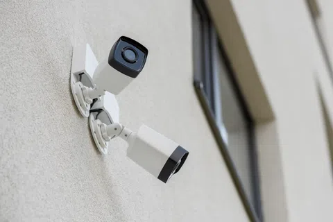 Two white security cameras mounted on a beige exterior wall near a window monitoring outside area.