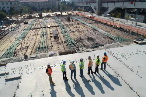 Construction workers in safety gear inspecting a large building foundation with exposed piping and rebar.
