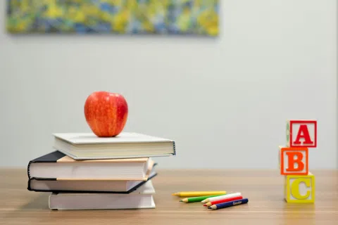 Stack of books with red apple on top, colorful pencils, and ABC blocks on wooden table in classroom setting