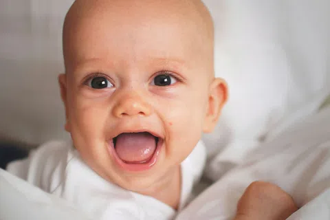 Happy baby with bright eyes and open mouth lying on white bed sheets and smiling joyfully