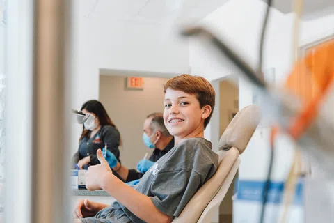 Teen boy smiling with braces, giving a thumbs up while sitting in a dental chair during a checkup.