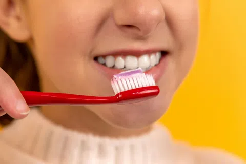 Person smiling with white teeth holding a red toothbrush with purple toothpaste on yellow background