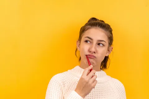 Thoughtful young woman in white sweater holding red pen near lips with bright yellow background.