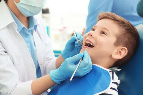 Child smiling while dentist wearing mask and gloves examines his teeth with dental tools.