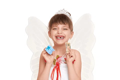 Smiling girl with missing teeth wearing fairy wings and holding a small gift box and money against white background
