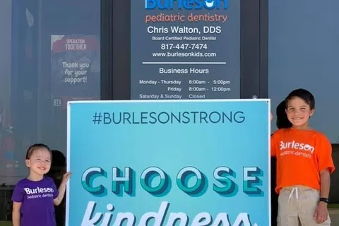 Two children stand beside a blue sign reading Choose kindness outside Burleson pediatric dentistry office.