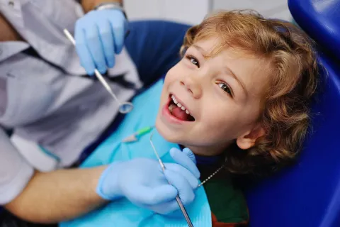 Smiling child with open mouth receiving dental checkup from dentist wearing blue gloves.