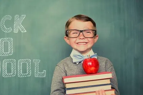 Smiling young boy with glasses holding books and an apple, standing in front of a chalkboard with Back to School text.
