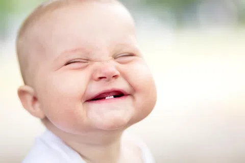 Close-up of a happy baby smiling with eyes closed showing two baby teeth, blurred background.