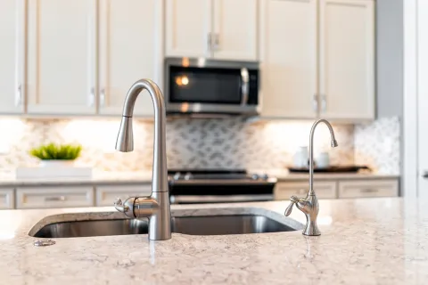 Modern kitchen sink with stainless steel faucets set in a granite countertop with white cabinetry and backsplash.