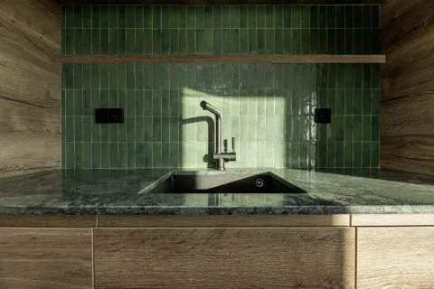 Modern kitchen sink with green tiled backsplash and wooden cabinetry under natural light.