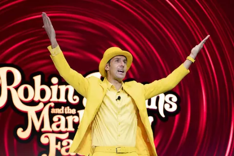 Man in bright yellow suit and hat gestures with arms raised on stage with red swirl backdrop and lettering.
