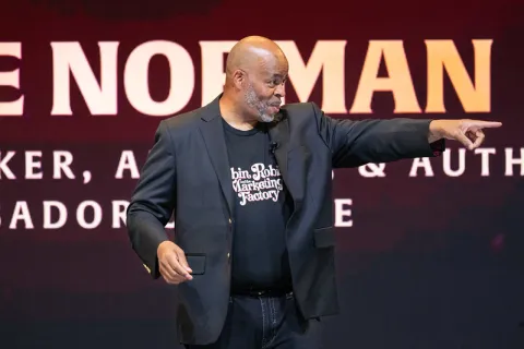 Speaker in black blazer and graphic tee gestures during presentation on stage with large text backdrop.
