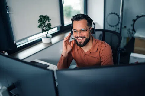 Smiling man wearing headset working at dual monitors in modern office with natural light and indoor plants