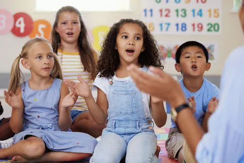 Diverse group of children sitting on a colorful mat singing and clapping in a classroom.