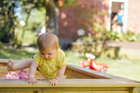 Toddler in a yellow striped shirt climbing out of a wooden sandbox in a sunny backyard with toys in the background