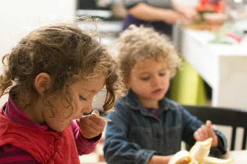 Two young children eating fruit at a wooden table while an adult prepares food in the kitchen background