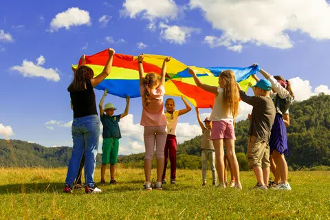 Group of children playing outdoors with a colorful parachute on a sunny day in a grassy field.