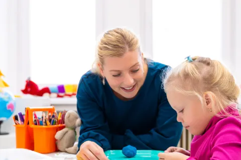 Teacher engaging with a young girl in a colorful classroom setting, playing with modeling clay and toys.