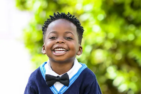 Smiling young boy in blue shorts, navy sweater with bow tie and blue shoes standing outdoors on paved surface.
