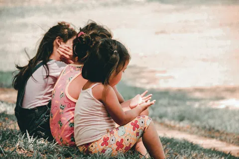 Three young girls sitting on grass outdoors enjoying a sunny day with casual summer clothing.