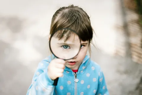 Young girl in blue polka dot jacket holding magnifying glass to her eye outdoors