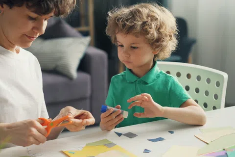 Adult and child crafting together using scissors and glue at a table with paper pieces in a cozy room.
