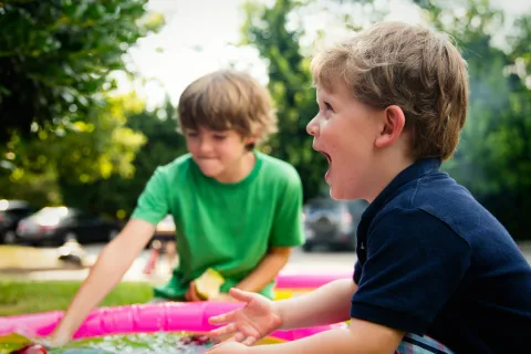Two young boys playing excitedly in a small pink inflatable pool outdoors on a sunny day