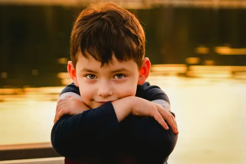 Portrait of a young boy with brown hair resting his arms and smiling softly by a glowing lakeside at sunset.