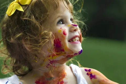 Smiling toddler girl with curly hair and yellow bow covered in colorful paint outdoors on a green background.