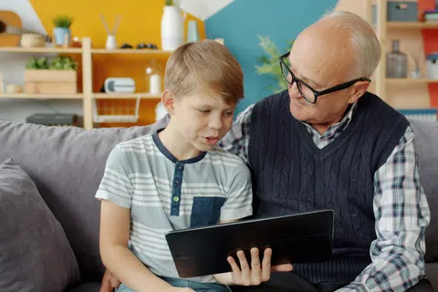 Grandfather and grandson sitting on couch using tablet together in colorful living room.