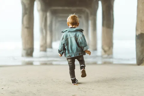 Young child wearing denim jacket walking on sandy beach under pier pillars toward ocean water.