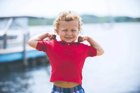 Smiling toddler boy flexing muscles in red shirt by the waterfront on a sunny day.