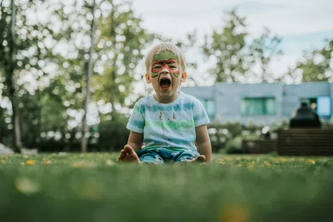 Young child with face paint sitting on grass yelling outdoors with trees and buildings in background.