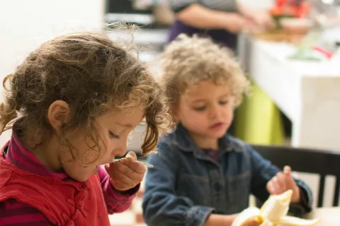 Two young children eating fruit at a wooden table in a cozy kitchen with a woman cooking in the background