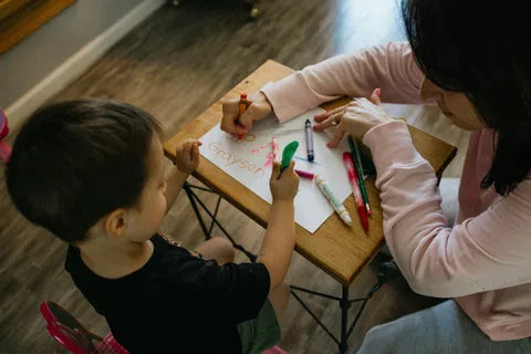 Child and adult drawing with crayons and markers on a small wooden table indoors