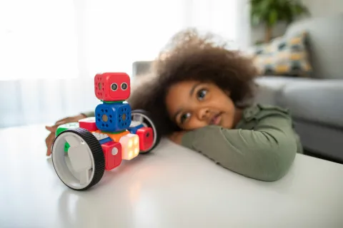 Curious child watches colorful robotic toy with large wheels and glowing lights on white table indoors.