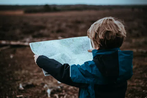 Child in blue jacket holding and reading a map outdoors in a natural, barren landscape.