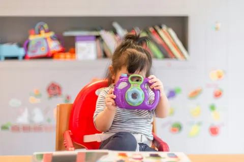 Young child sitting in a red high chair playing with a purple and green toy camera indoors.
