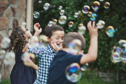 Children playing and chasing iridescent soap bubbles outdoors on a grassy backyard near a house.