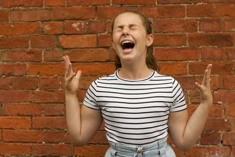 Young woman with eyes closed and hands raised, expressing frustration in front of a red brick wall.