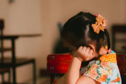 Young girl in a floral dress intently reading a colorful book while sitting at a wooden desk in a classroom.