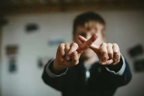 Close-up of a child crossing fingers to form an X sign with blurred background indoors.
