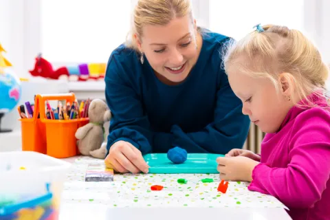 Teacher and child engaging in creative play with modeling clay and toys at a bright classroom table.